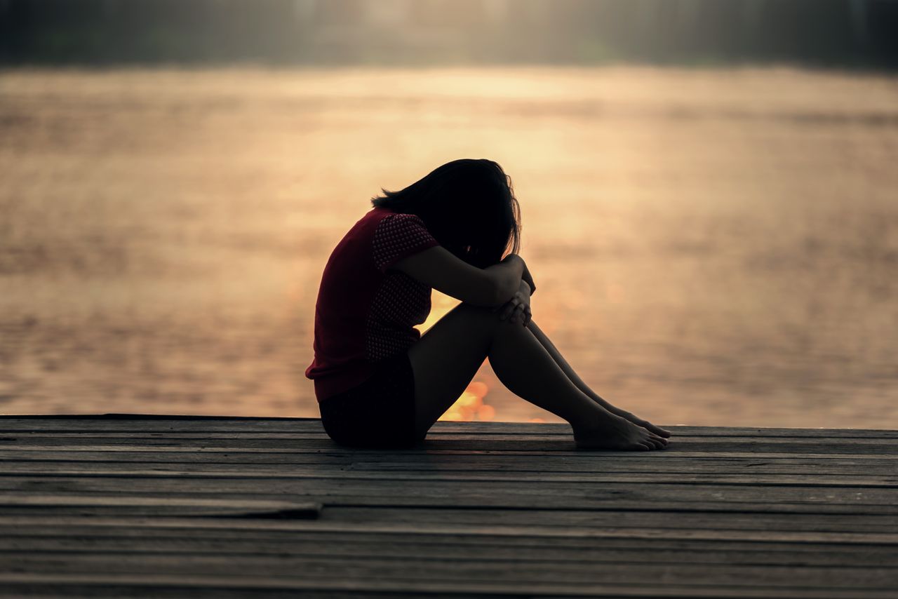 A silhouetted woman sits alone on a wooden dock at sunset, hugging her knees and looking down in a quiet, emotional moment.